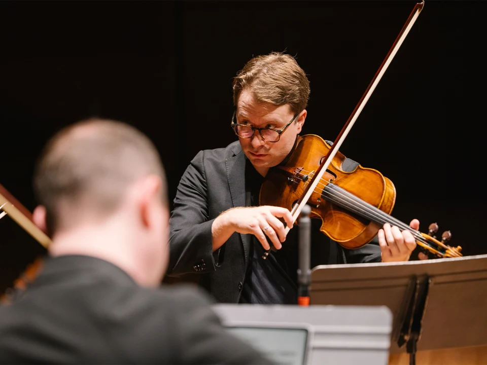 A focused violinist performs on stage, wearing a black suit and glasses. Another musician in the foreground appears to be playing or observing. Music stands are visible in the background.