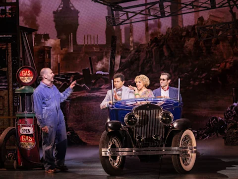 Four actors perform on stage; one stands by vintage gas pumps while three sit in a classic blue car with an industrial city backdrop behind them.