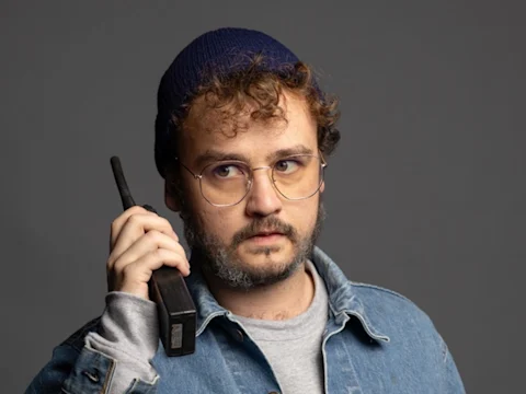 A person wearing glasses, a blue beanie, and a denim jacket holds a walkie-talkie to their ear, looking to the side against a plain gray background.