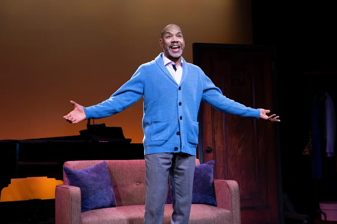 A man in a blue cardigan stands with arms open, smiling, in front of a sofa on a stage set with warm lighting.