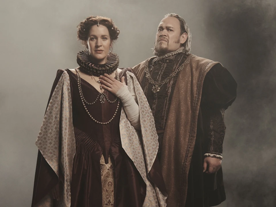 A woman in a Renaissance-style gown gazes at a man in period clothing with a heavy chain around his neck.