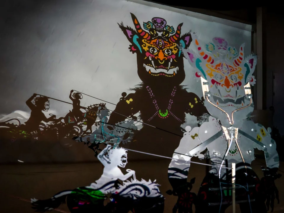 Colorful shadow puppets with intricate designs cast shadows on a backdrop, depicting mythical creatures in an illuminated setting.