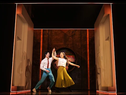 Two people dance energetically on stage between two modern door frames, with warm lighting and a textured, circular backdrop behind them.