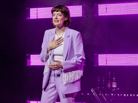 A person in a light purple suit with fringe stands on stage, holding a hand to their chest, with bright pink-purple LED lights in the background.