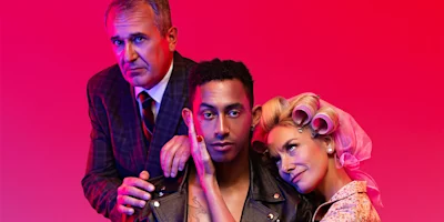 Three people pose against a red background; a man in a suit stands behind a younger man in a leather jacket, while a woman with curlers leans affectionately on the younger man's shoulder.