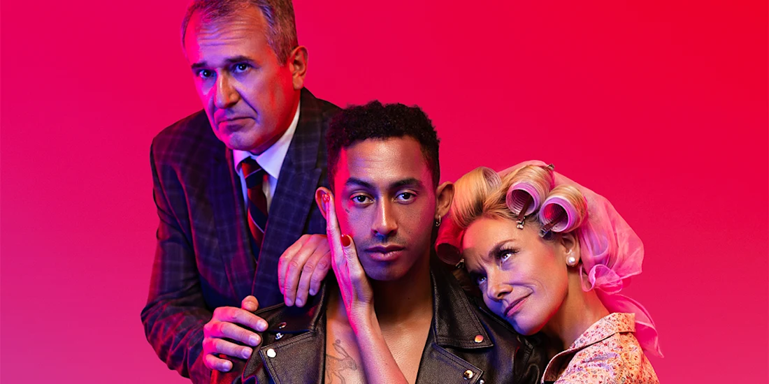 Three people pose against a red background; a man in a suit stands behind a younger man in a leather jacket, while a woman with curlers leans affectionately on the younger man's shoulder.