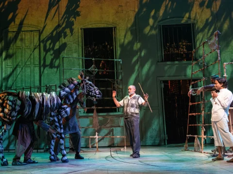 Group of actors on stage, two manipulating a large puppet resembling an animal, one with arms raised, and another dressed in white. Scenery includes a structure with bars and greenery projections.