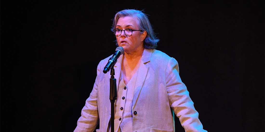 A person in glasses and a light suit speaks into a microphone on stage against a black background.
