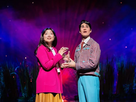 Two actors stand on stage in colorful costumes, holding a glass jar, with tall grass and dramatic purple lighting in the background.