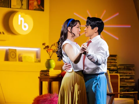 A man and woman in vintage-style clothing dance together in a warmly lit room with records, books, and a retro decor.