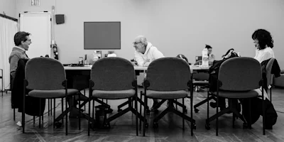 Three people sit at a rectangular table in a meeting room, facing each other, with empty chairs in the foreground. The setting appears casual and focused.