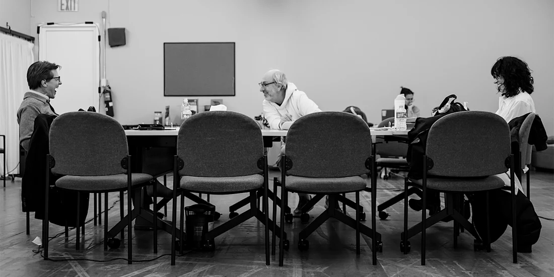 Three people sit at a rectangular table in a meeting room, facing each other, with empty chairs in the foreground. The setting appears casual and focused.