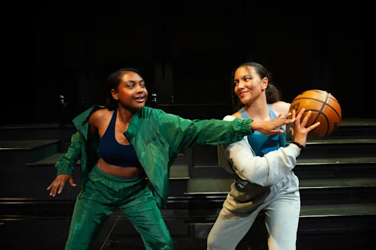 Two women in athletic wear play basketball indoors; one holds the ball while the other attempts to block or steal it.