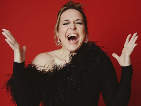 Woman wearing a black, one-shoulder, feathered dress smiles broadly with eyes closed and hands raised against a solid red background.
