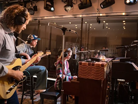 Three musicians wearing headphones play guitar, bass, and keyboard together in a recording studio, separated by a glass partition.