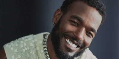 A man with a beard and short curly hair smiles at the camera, wearing a light-colored shirt with sequins and a chunky chain necklace.