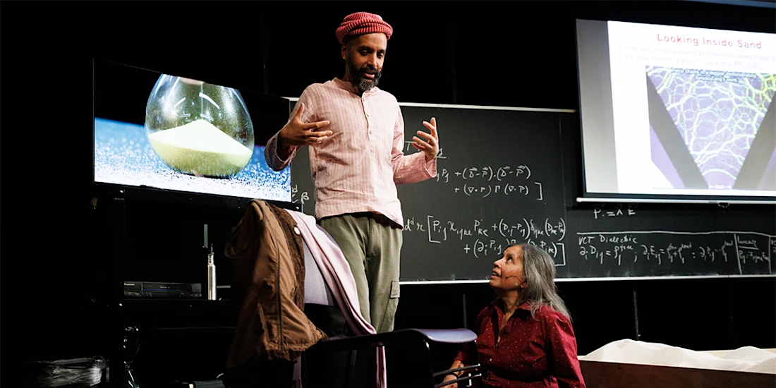 A man stands and speaks next to a chalkboard filled with equations, while a woman sits and looks up at him; scientific images are displayed on screens nearby.