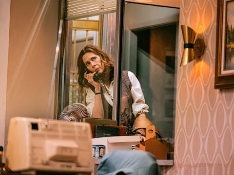A woman stands behind a glass partition, talking on a corded phone in an office with retro decor and patterned wallpaper.