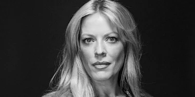 A woman with long, light hair looks directly at the camera against a dark background in a black and white portrait.