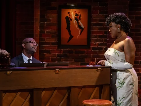 A man plays a piano while a woman in a floral dress stands nearby holding a drink. A framed picture hangs on a brick wall behind them.