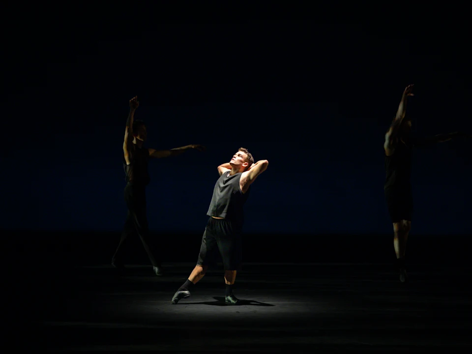 A dancer in dark clothing performs on a dimly lit stage, striking a pose with arms behind head. Two other dancers are visible in the background.