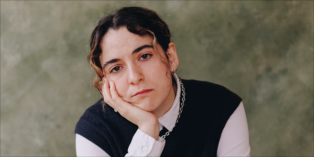 Person with curly hair rests their face on their hand, looking at the camera with a neutral expression. They wear a white shirt, dark vest, and chain necklace. Green textured background.