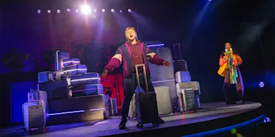 A performer sings on stage surrounded by stacked suitcases, while another person stands in the background under theatrical lighting.