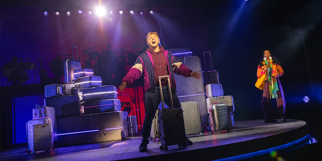 A performer sings on stage surrounded by stacked suitcases, while another person stands in the background under theatrical lighting.