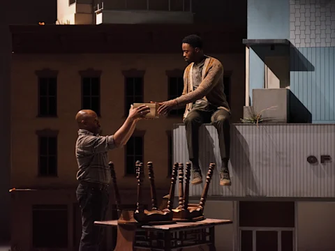 Two men interact on a stage set; one man sits on a building ledge reaching down, while the other stands below holding a cup, near a table with stacked chairs.