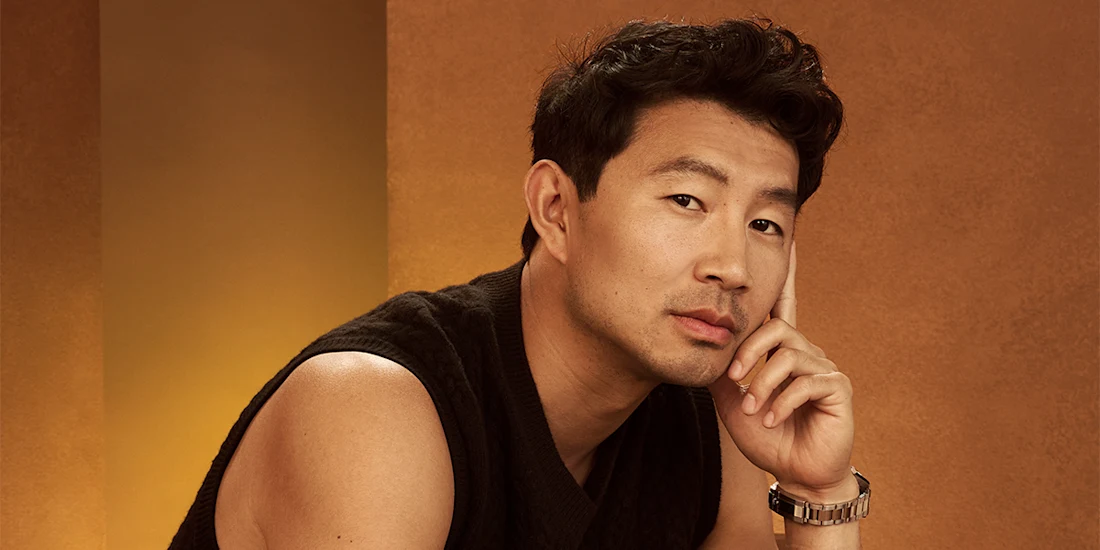 A man with short black hair wearing a sleeveless black sweater and a watch rests his chin on his hand, seated against a brown and gold background.