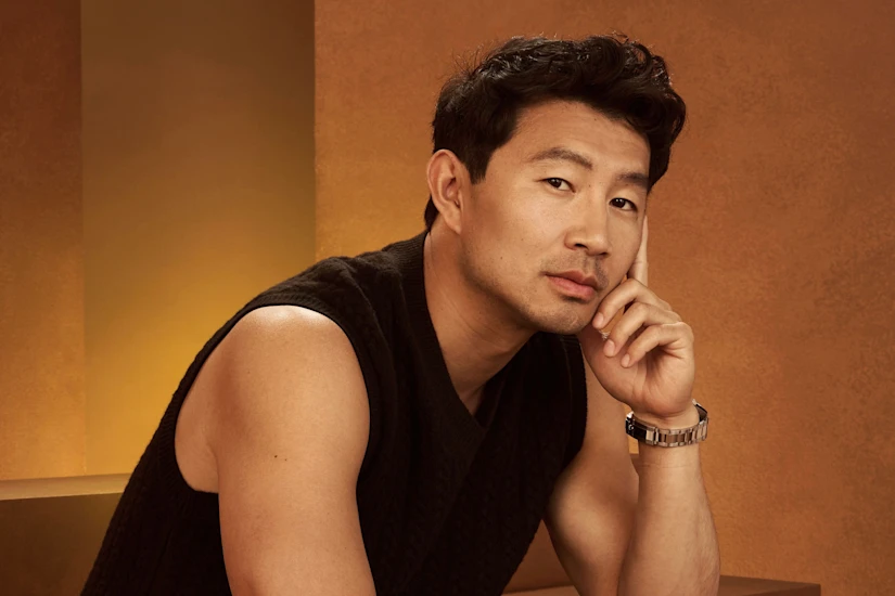 A man with short black hair wearing a sleeveless black sweater and a watch rests his chin on his hand, seated against a brown and gold background.