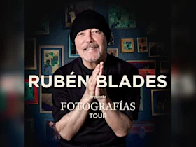 Rubén Blades smiles and clasps his hands in front of a wall with framed photos; text reads "Rubén Blades presenta Fotografías Tour.