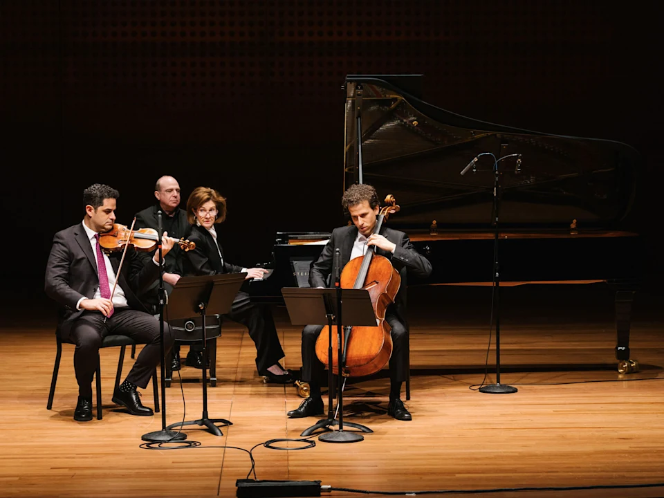 Four musicians perform on stage with a violin, cello, and piano; music stands and microphones are set up in front of them.