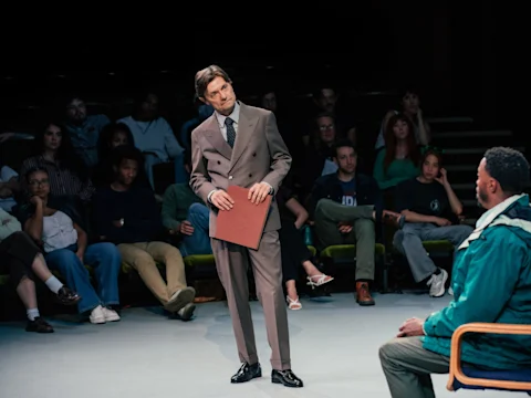 A man in a suit stands holding a folder, addressing another seated man, while an audience observes in a dimly lit room.