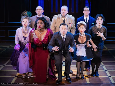 Eight actors in vintage costumes pose in a dramatic, crouched position on stage, appearing surprised or shocked, with a dark paneled backdrop behind them.