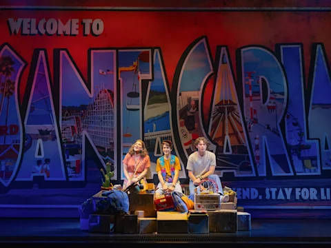 Three people sit on stage surrounded by props, in front of a large, colorful "Welcome to Santa Carla" mural with carnival and beach imagery in the background.