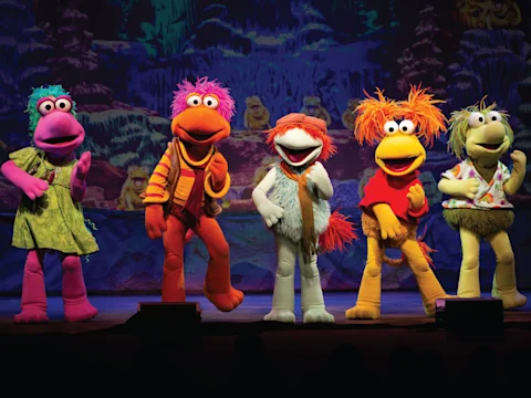 Five colorful puppet characters stand in a row onstage, posing energetically with a vibrant, illustrated backdrop behind them.