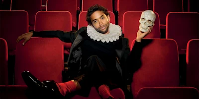 A person in a theater seat wears a ruffled collar and holds a skull, resembling a classic Hamlet scene, with red seats in the background.
