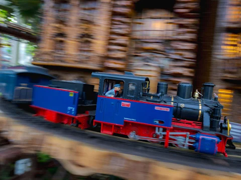 A blue and red miniature train with a driver moves quickly on an elevated track, with the background blurred to show motion.