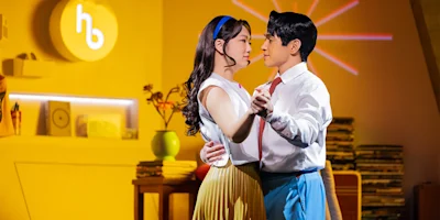 Two people wearing retro-style clothing dance closely in a brightly lit, vintage-themed room with records and decor in the background.