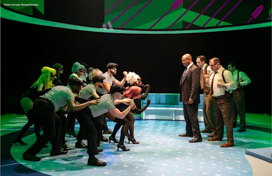 A group of performers in striped shirts face three people in suits and suspenders on a brightly lit stage with green and blue lighting effects.