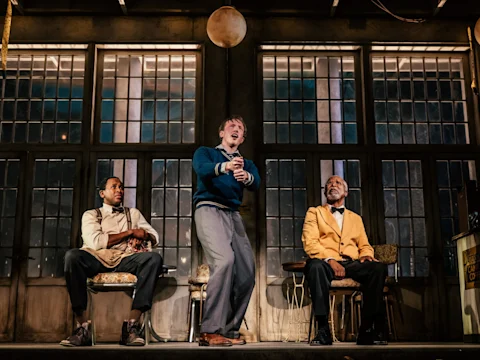 Three men sit on stage in front of large windows, with the man in the center standing and gesturing passionately, while the other two watch him.