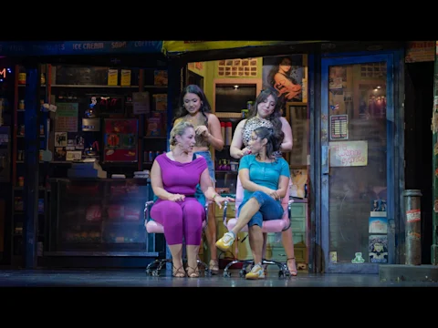 Four women sit and stand in a brightly lit salon setting on stage, two seated in pink chairs talking, while two stand behind them, with shelves and signage visible in the background.