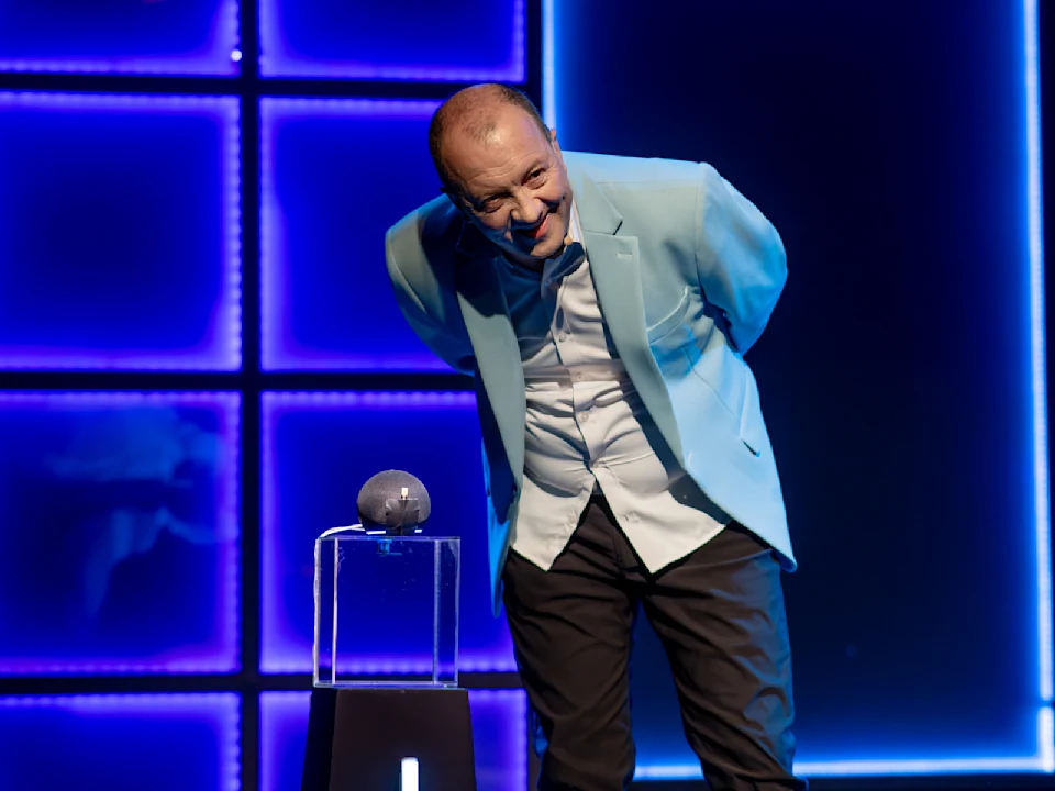 A man in a light blue blazer stands on stage, leaning forward with hands behind his back, next to a glass stand displaying a small round object under blue lighting.