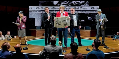 Five actors perform on stage, one holding a large newspaper, with an audience surrounding them and a backdrop featuring The New York Times headlines.