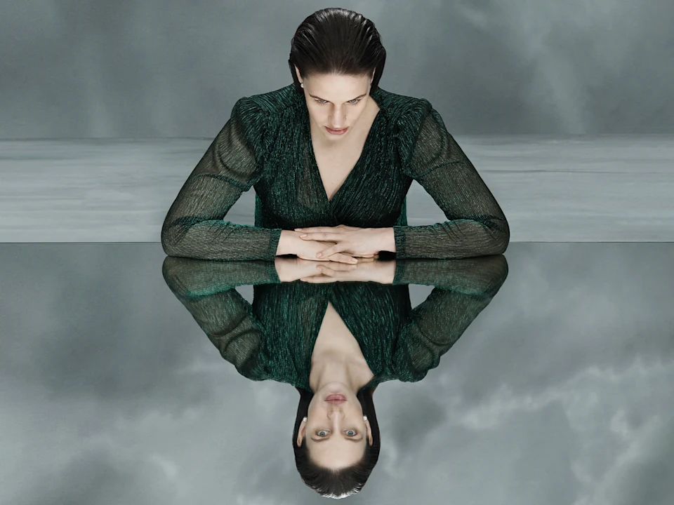 A woman in a green dress sits at a reflective surface, looking down at her own mirrored image, with a grey, cloudy background.