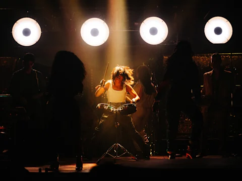 A drummer performs energetically on stage under bright lights, surrounded by band members and silhouetted dancers.