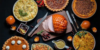 A table set with a glazed ham, various pies, mashed potatoes, green bean casserole, dinner rolls, cranberry sauce, and gravy on a dark background.