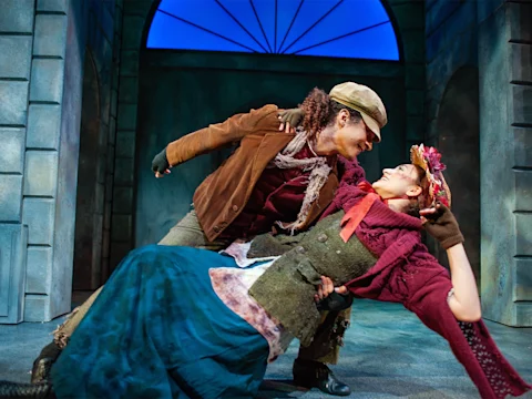 Two actors in period costumes perform a dramatic dance pose on stage, with a blue-lit backdrop suggesting an outdoor setting.