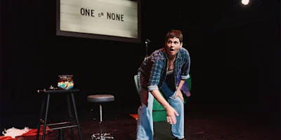 A person performs on stage in front of a sign reading "One on None," with a microphone, stool, and a bowl on a table nearby.